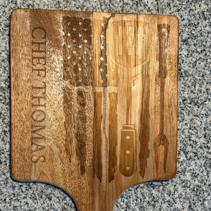 Wooden cutting board with engraved American flag and 'Chef Thomas' text on a gray surface